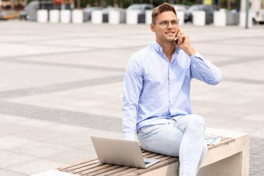 Dışarıda bankta cep telefonuyla konuşan dizüstü bilgisayarı olan yakışıklı bir genç.