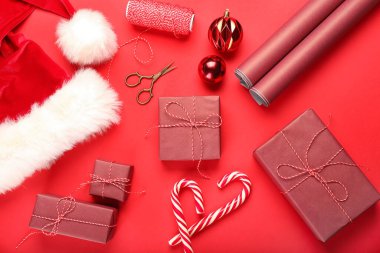 Hediye kutuları, ambalaj kağıtları ve kırmızı arka planda Noel Baba şapkası olan kompozisyon