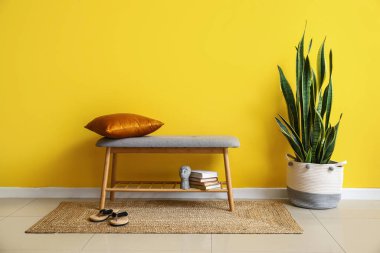 Yastıklı, kitaplı, parmak arası terlikli ve sarı duvarlı rahat bir bank.