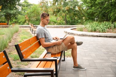 Parktaki bankta dizüstü bilgisayar kullanan güneş gözlüklü yakışıklı genç adam.