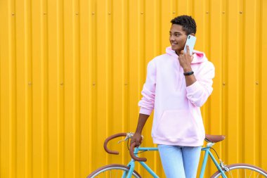 Bisikletli Afro-Amerikalı genç çocuk sarı çitin yanında cep telefonuyla konuşuyor.