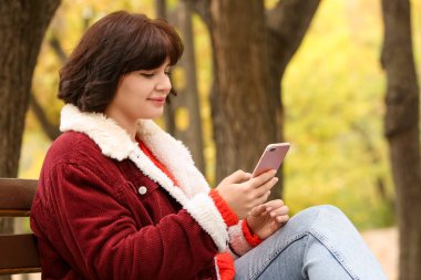 Güz parkında bankta oturan ve cep telefonu kullanan güzel bir kadın.