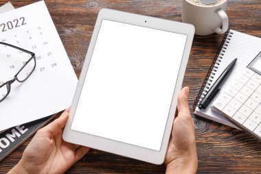 Tablet bilgisayarı olan kadın eller, 2022 yılı için kağıt takvim ve ahşap arka planda kırtasiye malzemeleri.