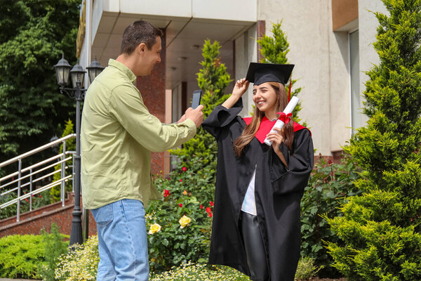 Happy man taking photo of his daughter in graduation gown and with diploma outdoors