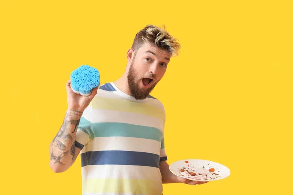 Überraschter Junger Mann Mit Schwamm Und Schmutzigem Teller Auf Gelbem — Stockfoto