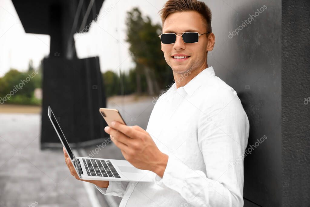 Young man in sunglasses with laptop using mobile phone outdoors