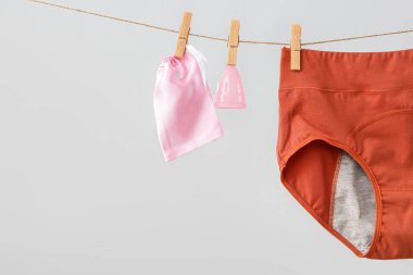 Adet külotu, adet kupası ve çanta gri arka planda asılı duruyor, yakın plan.