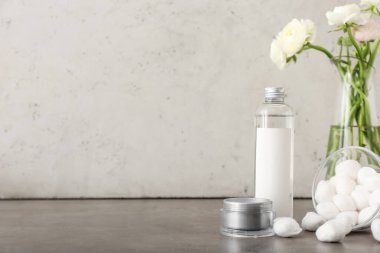 Kozmetiklerle dolu bir şişe, yüz kremiyle dolu bir kavanoz ve gri masadaki ranunculus çiçekli vazo.