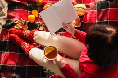 Elinde çay fincanı olan bir kadın ekose üzerinde oturuyor ve elinde boş sayfalarla kitap tutuyor.