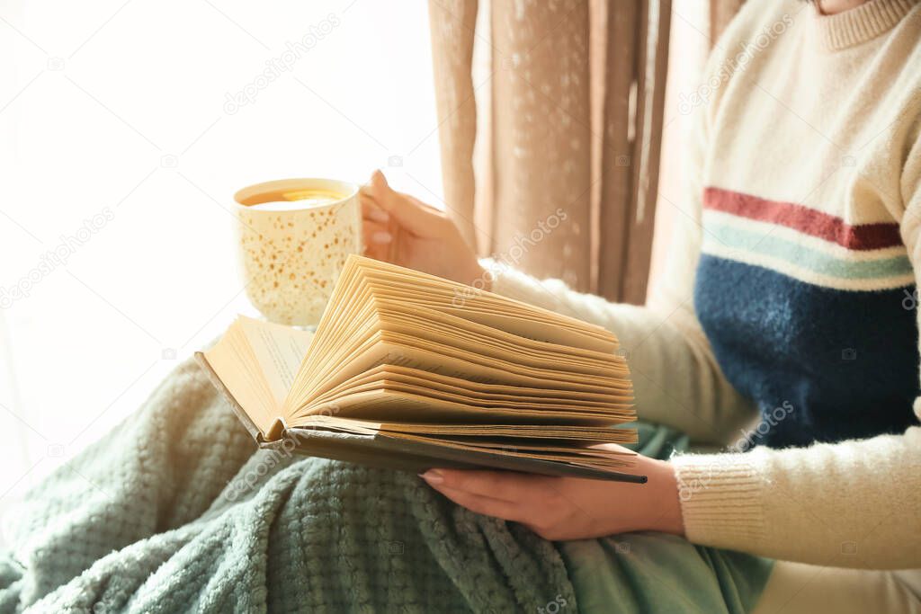 Woman with plaid holding cup of tea and reading book in room