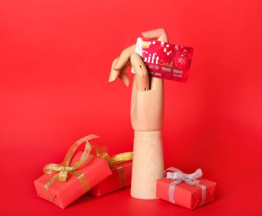 Tahta el, hediye kartı ve kırmızı arka planda hediyeler.