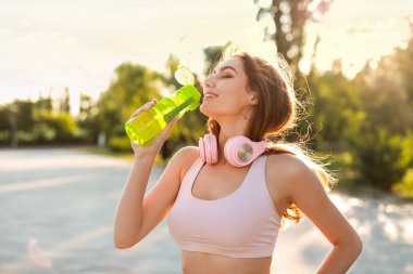 Kulaklıklı genç ve güzel bir kadın. Açık hava eğitiminden sonra su içiyor.