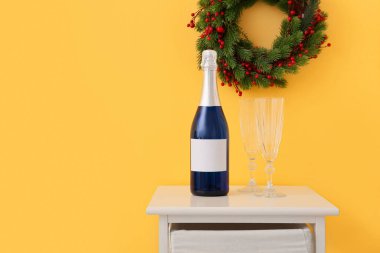 Bir şişe şampanya, masada bardaklar ve renkli duvarda Noel çelengi