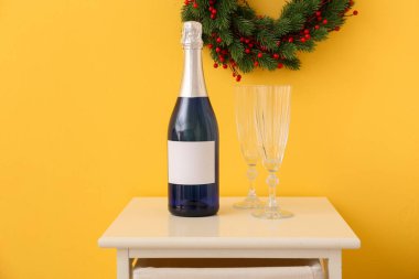 Bir şişe şampanya, masada bardaklar ve renkli duvarda Noel çelengi