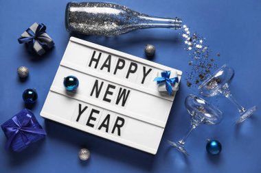 Mutlu yıllar yazılı pano, şampanya şişesi, gözlük ve mavi arka planda Noel dekoru.