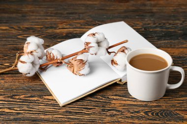 Bir fincan lezzetli kahve, boş kitap ve tahta arka planda pamuk çiçekleri.