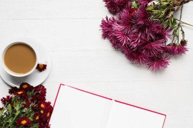 Bir fincan lezzetli kahve, boş kitap ve beyaz ahşap arka planda kasımpatı çiçekleri.