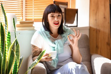 Dövmeli genç bir kadın kahve içiyor ve evde görüntülü sohbet ediyor.