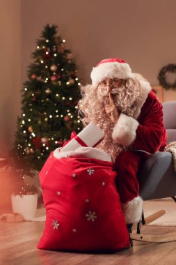 Noel Baba çantasıyla Noel arifesinde odada sessizlik işareti yapıyor.