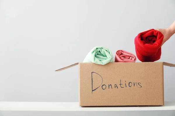 Woman putting donation clothes in box on shelf against light background