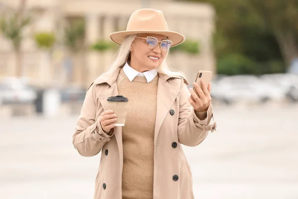 时尚的成熟女人 头戴时髦的毛毡帽 城市街道上端着纸杯咖啡和手机 — 图库照片
