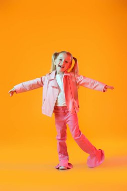 Pembe ceketli şirin küçük kız turuncu arka planda dans ediyor.