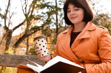 Güz parkında elinde bardakla içki içip kitap okuyan genç bir kadın.