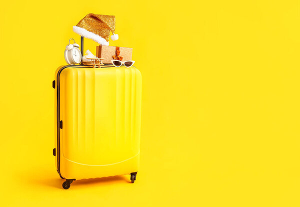 Suitcase with sunglasses, Christmas gift box, wooden airplane, alarm clock and Santa hat on yellow background