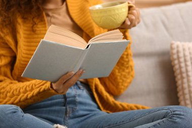 Evdeki kanepede bir fincan sıcak çay içip kitap okuyan genç bir kadın.