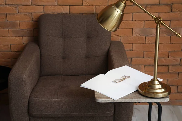 Oturma odasındaki koltuğun yanındaki sehpanın üzerinde parlayan lamba, kitap ve gözlük.