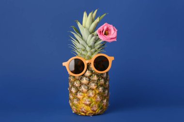 Mavi arka planda çiçek olan güneş gözlüklü komik ananas.