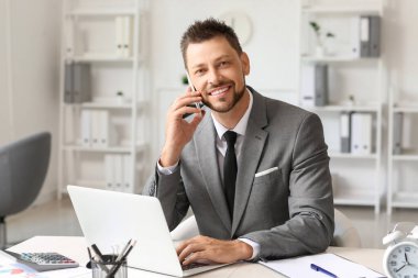 Ofiste cep telefonuyla konuşan dizüstü bilgisayarlı gülümseyen bir işadamı.