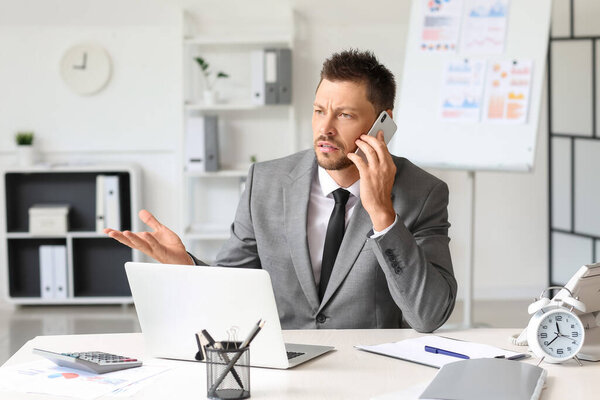 Upset young businessman talking by mobile phone at table in office