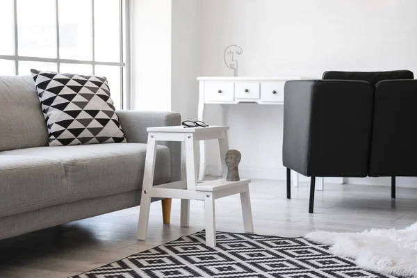 Oturma odasındaki kanepenin yanında gözlüklü ve dekorlu beyaz bir tabure.