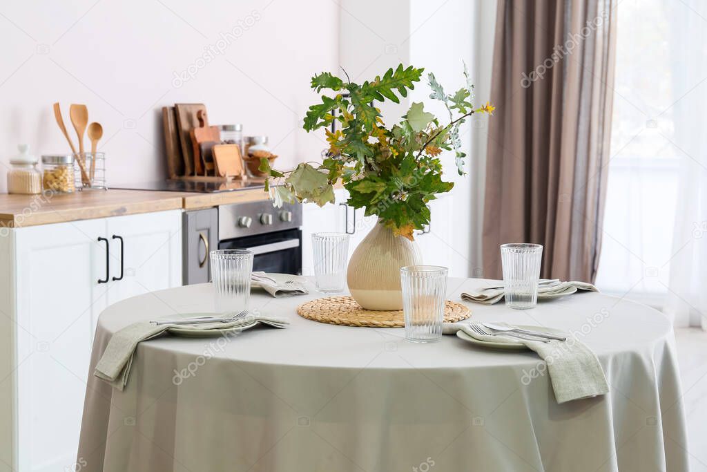 Dining table with setting and vase with autumn branches in modern kitchen