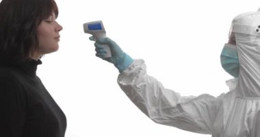 Koruyucu kıyafetli doktor beyaz arka planda kızılötesi termometresi olan bir kadının sıcaklığını ölçüyor.