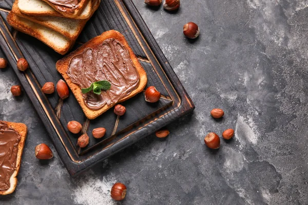 Wooden board of bread with chocolate paste and hazelnuts on black background, closeup