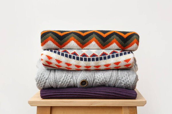 Stack of stylish sweaters on wooden stool against light background