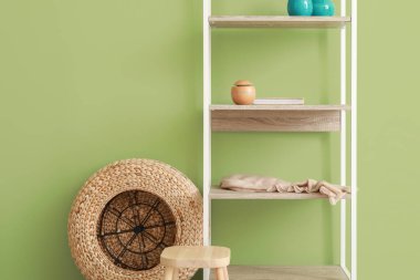 Dekorlu raf ünitesi, rattan puf ve yeşil duvarın yanındaki tabure.