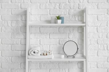 Beyaz tuğla duvarın yanında banyo aksesuarları olan raf ünitesi.