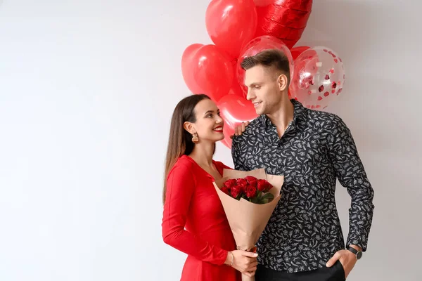 Happy young couple with bouquet of flowers and balloons on light background. Valentine's Day celebration