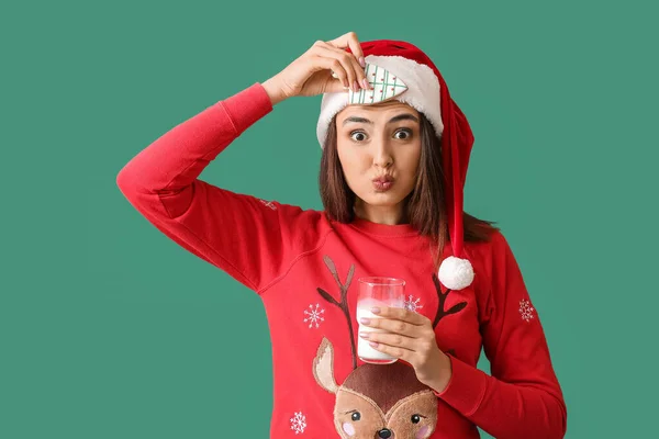 面白いです若いです女性でサンタ帽子とともにジンジャーブレッドクッキーとガラスのミルクの緑の背景 — ストック写真