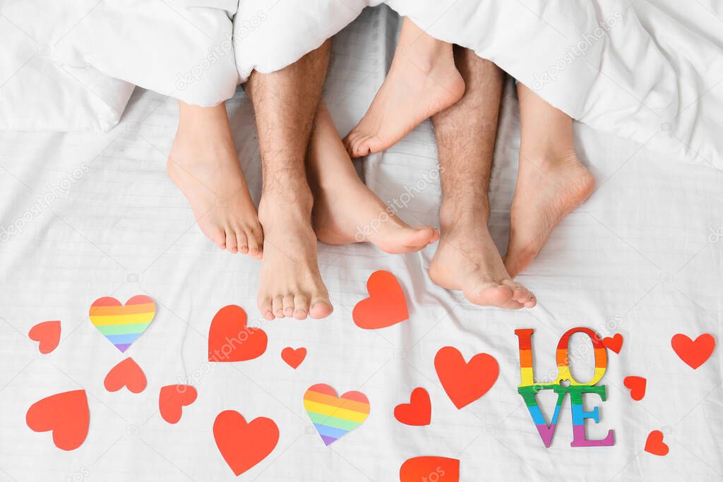 Legs of young man and two women lying in bed covered with paper hearts, top view. Concept of polyamory and LGBT