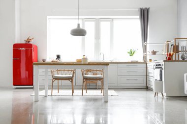 Kırmızı buzdolabı, beyaz tezgah ve yemek masası olan aydınlık modern mutfak.