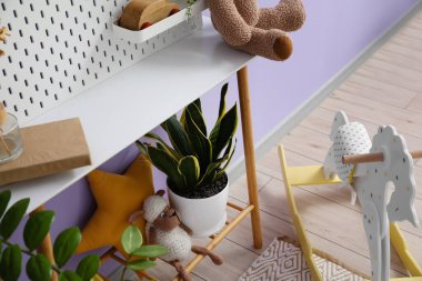 Tahtalı masa, ev bitkisi, oyuncak ve yastık, leylak duvarın yanında.