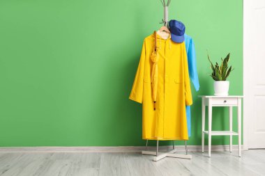 Yeşil duvarın yanında yağmurluk, şemsiye, şapka ve masa var.