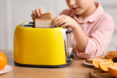 Küçük çocuk mutfakta ekmek kızartma makinesine ekmek dilimleri koyuyor.