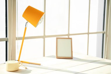 Modern lamba, bardak ve boş fotoğraf çerçevesi büyük pencerenin yanındaki beyaz ahşap masada.