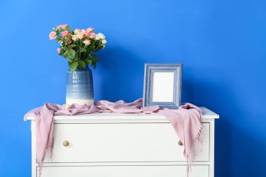 Mavi duvarın yanındaki çekmecelerde güzel güller, çerçeve ve pembe eşarplı vazo.
