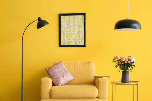 Vase with bouquet of beautiful fresh roses on table and comfortable armchair near yellow wall